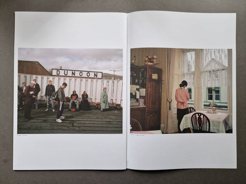An open publication laid flat on a dark surface, showing two printed photographs side by side. The left page contains a photo of people standing outdoors near a white sign reading DUNOON, and the right page contains a photo of a woman standing in a room by large windows.