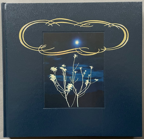 A square hardcover book or album with a textured dark blue cover, featuring a central window-like inset showing a night sky scene with a bright moon and white flowering branches, plus a decorative gold ornamental frame above. The item is photographed from above on a plain light gray surface.