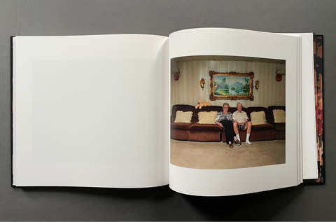 An open hardcover book laid flat on a dark gray background, showing a large blank white page on the left and a printed photograph on the right of two people sitting on a brown sofa in a living room.