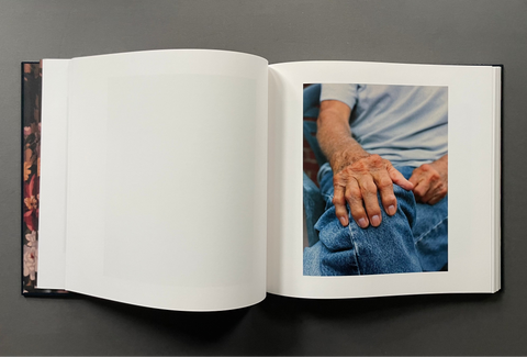 An open hardcover book laid flat on a dark gray surface, showing a large blank white page on the left and a printed photograph of a person's hands resting on blue jeans on the right.