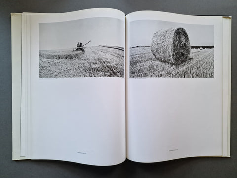 An open book spread showing two black-and-white photographs of a field scene: a combine harvesting on the left page and a large hay bale on the right page, photographed against a dark gray background.