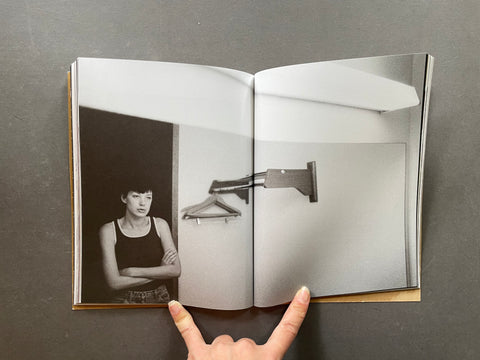 An open book or magazine lying on a dark gray surface, showing a black-and-white interior photograph spread across both pages. A person's hand is visible holding the bottom edge of the book.