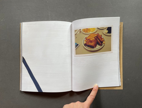 An open book or magazine lying on a dark gray surface, showing a printed photograph of a plate of food on the right page. A person's finger is pointing at the page.