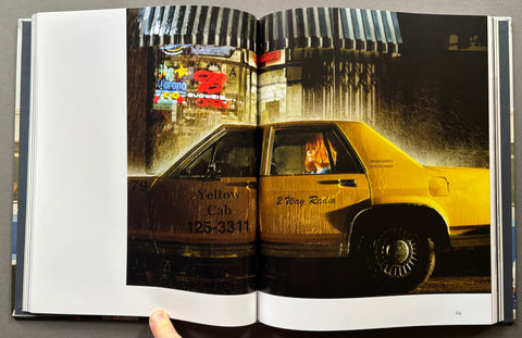 An open book showing a large color photograph printed on the pages, depicting a yellow cab in a rainy city street scene at night. A hand is visible holding the open book.