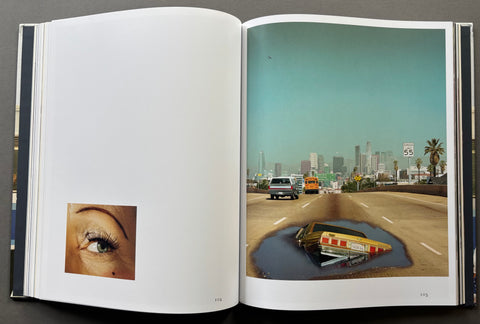 An open art book spread showing a surreal photograph on the right page with a city skyline, highway, and a car sinking into a puddle, alongside a close-up eye inset on the left page.