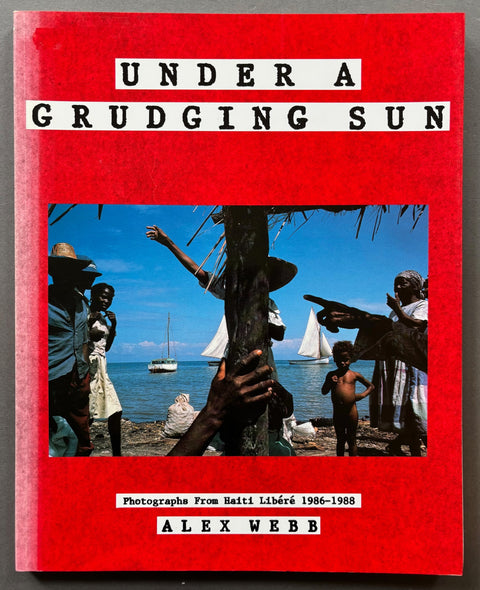 Book titled 'Under a Grudging Sun' by Alex Webb with a red cover featuring a photograph of Haitian people on the beach.