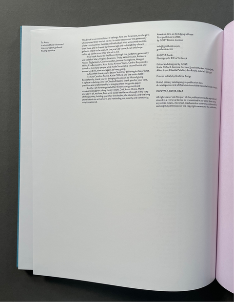 An open book laid flat on a dark gray surface, showing the interior title/plate page with dense black text on white paper and a pale pink page at the right edge.
