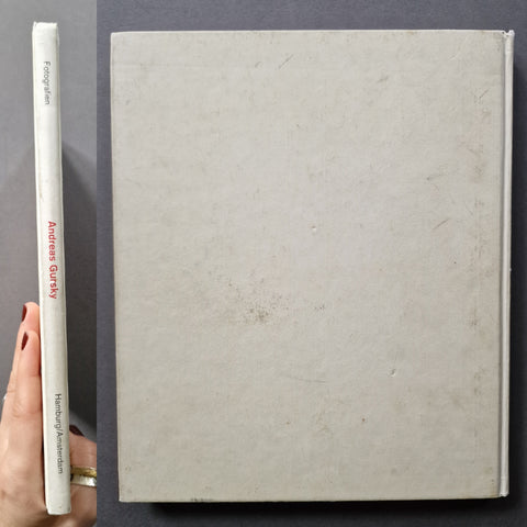 A hand holds a white hardcover book spine next to the full front cover of the same book, which has a textured off-white cover and shows the title 'Andreas Gribler' and 'History of Photography' in red on the spine.