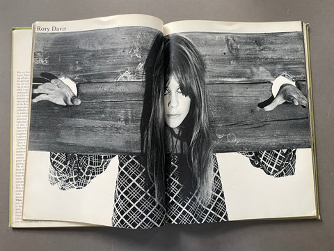 An open book showing a black-and-white printed photograph of a woman behind a wooden plank, with her hands extended outward. The book pages are spread open on a dark gray surface.