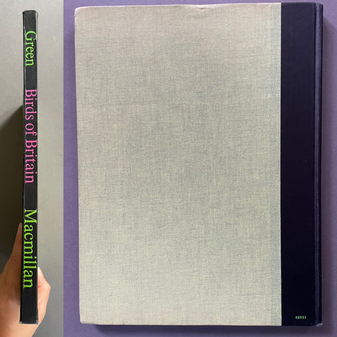 A hardcover book titled 'Green Birds of Britain' by Macmillan, held by a hand against a purple background. The book features a light gray linen-textured front cover, a black spine with colorful title text, and a black binding strip on the right side.