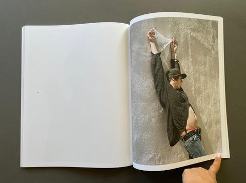 An open book or magazine lies flat on a dark surface, showing a printed fashion photograph on the right page of a person in a hat and shirt against a textured gray wall, with a hand holding the lower right edge of the page.
