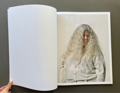 An open book spread against a dark gray background, showing a printed portrait of a person with very long, wavy white hair wearing a light-colored cardigan. A hand is visible holding the left page open.