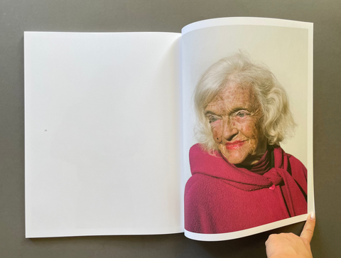 An open book lying on a dark gray surface, showing a printed portrait of an elderly woman with white hair wearing a red top on the right page. A hand is holding the lower right edge of the page.