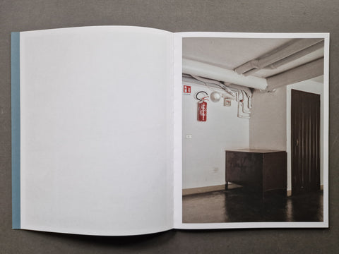 An open book, showing a large blank white page on the left and a printed interior scene on the right with a room, exposed ceiling pipes, a red fire extinguisher, and a dark cabinet.