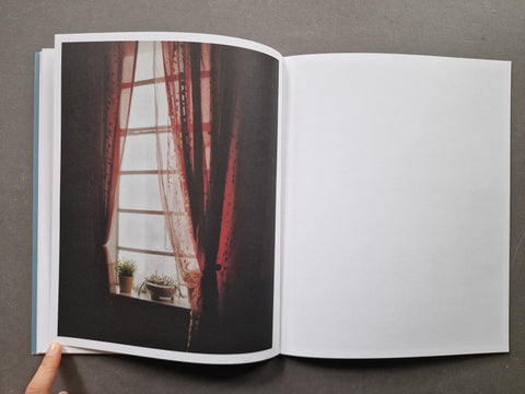 An open book, showing a printed photograph of red curtains by a window on the left page and a blank white page on the right. 