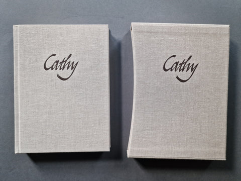 The linen book cover and its matching slipcase side by side on a dark gray background, both featuring the name 'Cathy' printed in black cursive script on the front cover.