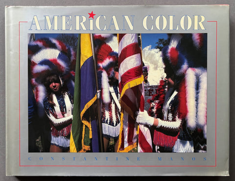 Book titled @American Color' by Constantine Manos with a grey cover featuring an image of people wearing headdresses and carrying flags.