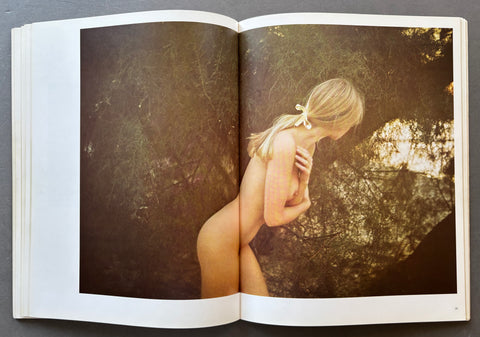 An open hardcover art book showing a large photographic print on the spread. The print features a sepia-toned nude woman with blonde hair tied back with a bow, set against a dark, textured background.