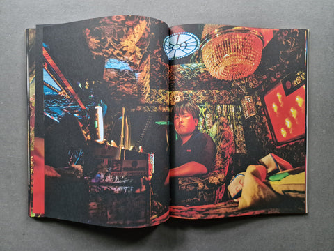 An open book. The pages contain a highly detailed, colorful, and cluttered interior scene with warm lighting, a hanging chandelier, screens, and a seated person visible in the printed photograph.