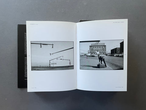 An open book or magazine spread laid flat on a dark gray surface, showing two black-and-white photographic prints on white pages. The left page contains a minimalist street scene with traffic lights, and the right page shows a person standing on a street corner in front of a building.