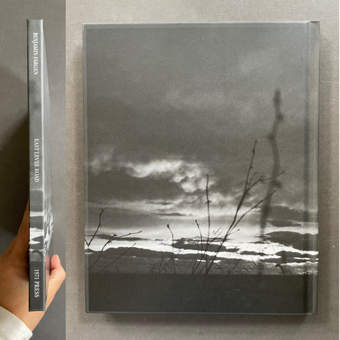 A hardcover book or art print with a black-and-white landscape photograph on the cover, shown standing upright next to its spine. A hand is holding the spine at the left edge.