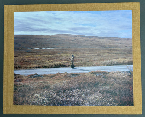 Book titled 'The Plateau' by Eirik Brekke with a brown cover featuring the image of a man on an electric unicycle travelling along a road surrounded by fields of heather.