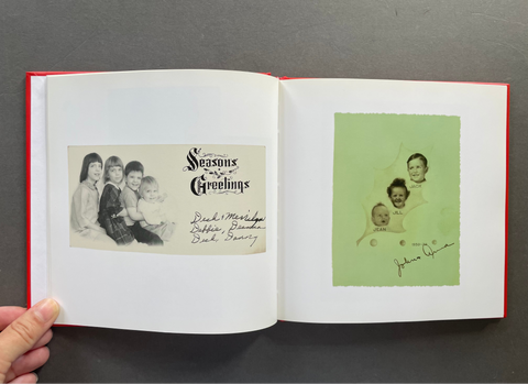 An open vintage photo album with a red hardcover, held open by a hand. The left page shows a black-and-white family portrait and a 'Seasons Greetings' card with handwritten signatures.
