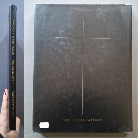 A hardcover book titled 'Gods of Earth and Heaven' by Joel-Peter Witkin, shown standing upright next to its front cover. A hand with painted nails is visible holding the spine on the left.