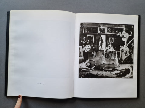 An open art book , showing a black-and-white photographic print on the right page and blank white paper on the left page. A hand with red nail polish is visible at the bottom left holding the book open.