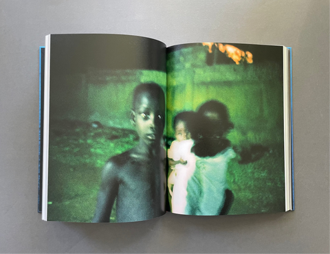 An open hardcover book lying flat on a neutral gray surface, displaying a double-page spread with a striking green-tinted photograph of a child on the left and a figure holding a baby on the right.
