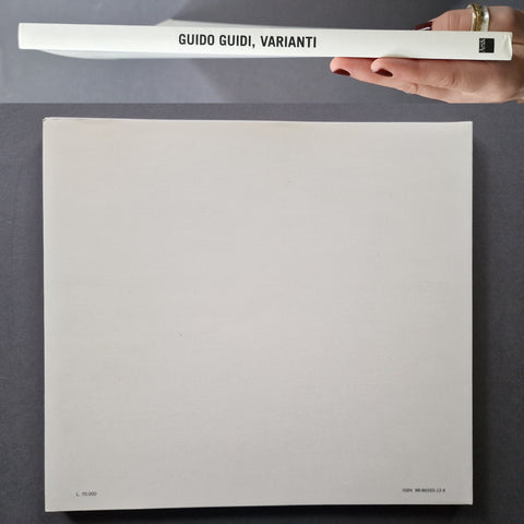 A hand holds a slim white book or booklet labeled 'GUIDO GUIDI, VARIANTI' above a large blank white sheet on a dark gray surface.