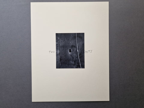 An original black and white silver gelatin art print  mounted on a large off-white sheet. The print shows a dark textured square with a small rectangular opening and handwritten text beside it reading 'TWO' and 'SHITS'.
