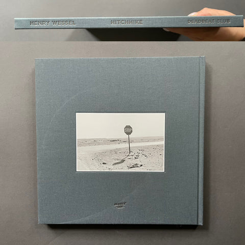 A hardcover book titled 'Henry Wessel: Hitchhike' is shown lying on a dark gray surface, with a hand holding the top edge. The cover is a muted blue-gray with a central black-and-white photograph of a desert road sign.