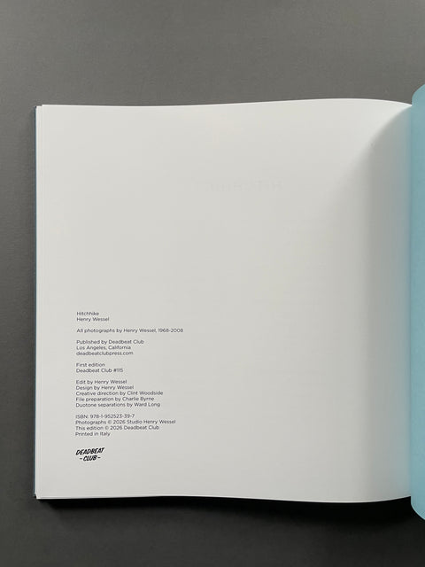 An open book or art print lying flat on a dark gray surface, showing a blank white page with small printed text on the left side and a light blue page or edge on the right.
