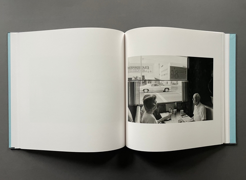 An open hardcover book laid flat on a dark gray background, showing a large blank white left page and a black-and-white photograph on the right page depicting two people seated at a table by a window.
