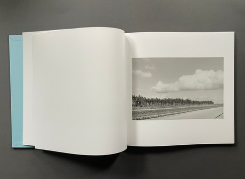 An open hardcover book or photo album laid flat on a dark gray surface, showing a black-and-white landscape photograph printed on the right page and a blank white left page. A light blue interior or backing sheet is visible along the left edge.