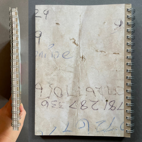 A spiral-bound notebook with a beige, stained cover featuring handwritten numbers and text, shown next to a hand holding a matching spiral-bound metal rod for scale, against a dark gray background.