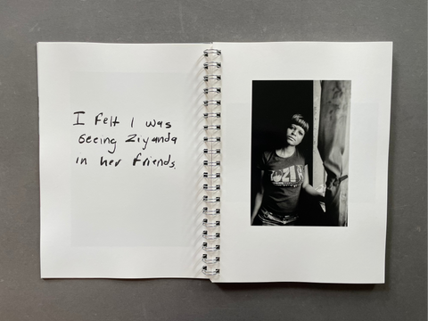 A spiral-bound notebook or journal lying open on a dark gray surface, showing handwritten text on the left page and a black-and-white photograph printed on the right page.