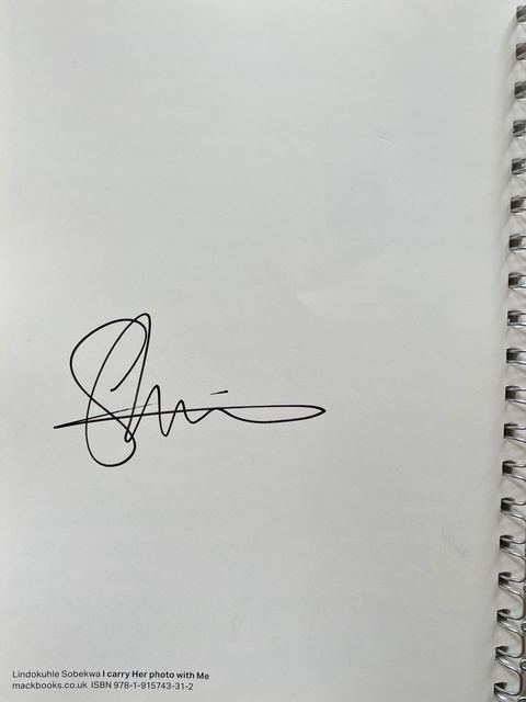 A white spiral-bound notebook or journal lying on a dark gray surface, featuring a black cursive signature printed on the cover and small text at the bottom left.