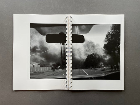 A spiral-bound photo print or notebook lies open on a dark gray surface. The center page shows a black-and-white street scene viewed from inside a car, with a rearview mirror, road, trees, and dark sky.