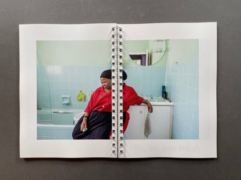 A spiral-bound photo print or notebook page displayed flat on a dark gray surface, featuring a photograph of a woman in a red top and dark skirt sitting in a tiled bathroom.