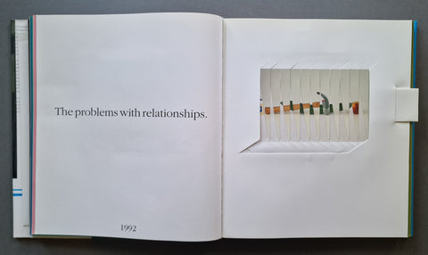 An open art book showing a printed page with the text 'The problems with relationships.' and a sculptural paper cutout on the right page depicting a small figure.