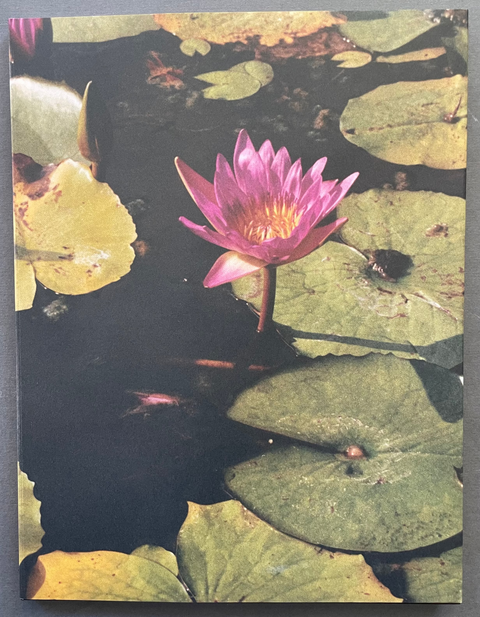 A rectangular art print or book cover featuring a pink water lily among green lily pads on dark water, photographed against a dark gray background.