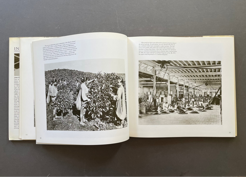 An open hardcover book laid flat on a dark gray surface, showing two printed pages with black-and-white photographs and text. The left page contains a photo of people working among plants, and the right page shows a large interior scene with many people seated or standing in a spacious hall.