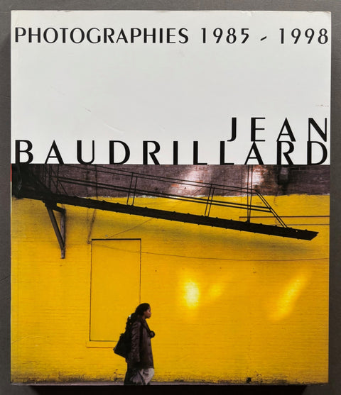Book titled 'Photographies 1985-1998' by Jean Baudrillard with a cover featuring the image of a woman walking under a fire escape.
