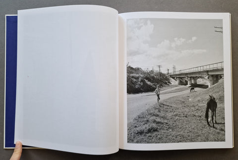 An open book, showing a mostly blank white left page and a black-and-white landscape photograph on the right page with a bridge, grassy hillside, and two horses and a person.