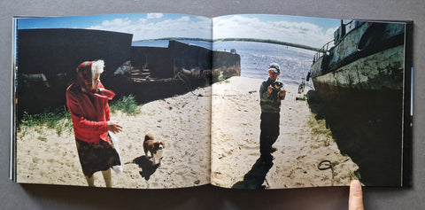 An open book, showing a two-page spread with a photograph of a beach scene featuring an old lady in a red jacket, a dog and a kid between two boats, and water in the distance.