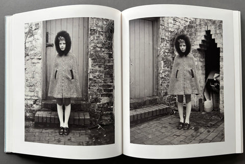 An open book displaying two black-and-white photographs side by side, each showing a young girl standing outdoors in front of a rustic wooden door and brick wall.