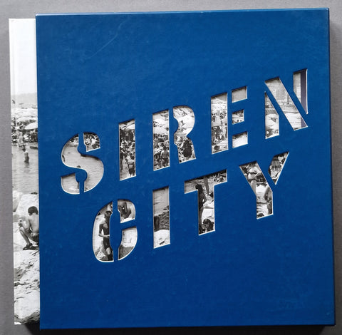 A square blue book cover titled 'SIREN CITY' with cutout lettering revealing black-and-white photographs, photographed against a neutral gray background.