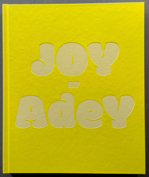 A bright yellow hardcover book with large, raised, cream-colored bubble letters spelling 'JOY by Adey' on the front cover, photographed against a dark gray background.